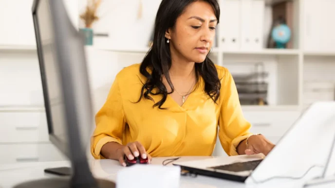 Woman in a yellow blouse sits at a white desk, using a computer mouse while working on a tablet in an office setting.
