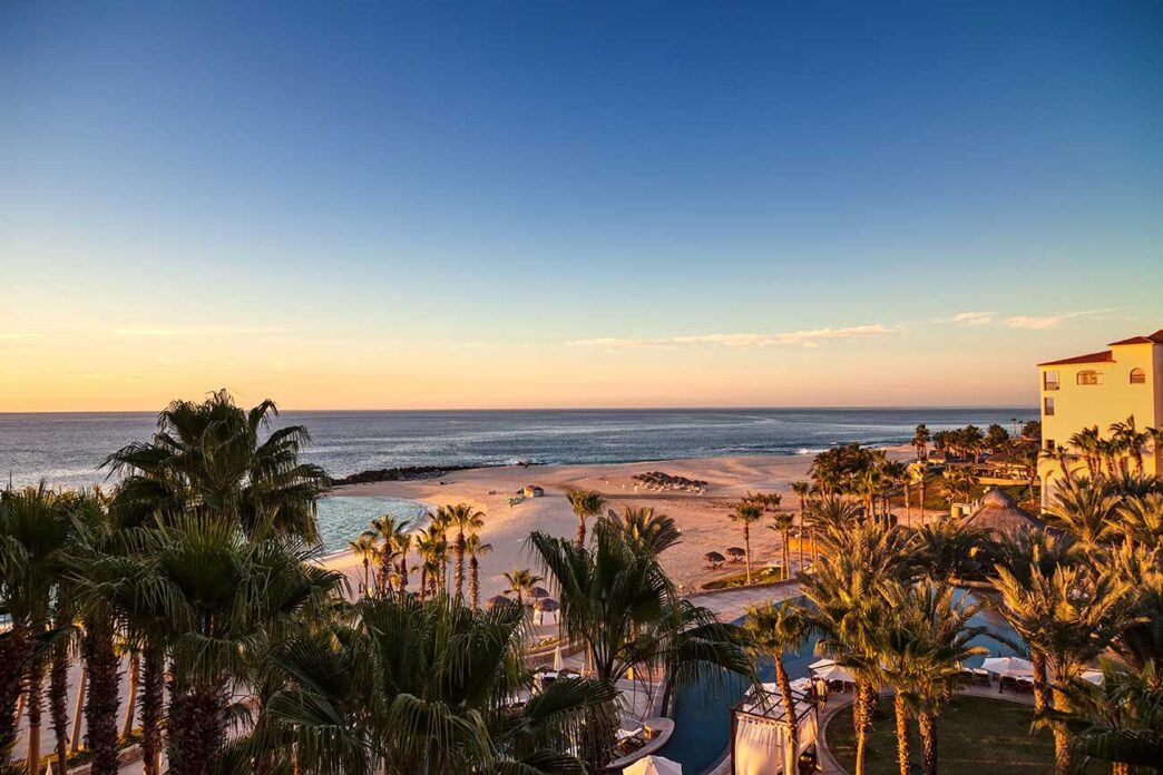 Sunset over a tropical beach with palm trees, a pool area in the foreground, and a calm sea beyond.