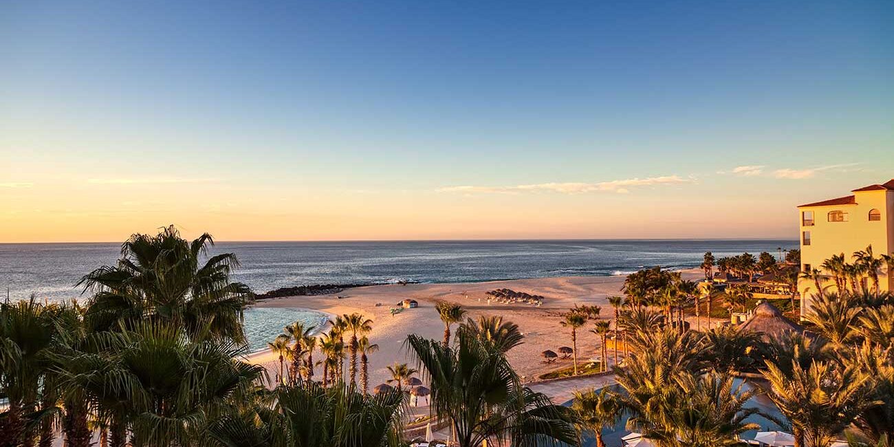 caboSunrise - Gringo Gazette Sunset over a tropical beach with palm trees, a pool area in the foreground, and a calm sea beyond.