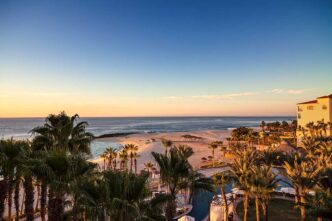 Sunset over a tropical beach with palm trees, a pool area in the foreground, and a calm sea beyond.