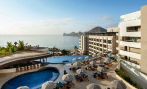 Resort pool area with numerous lounge chairs and striped umbrellas, ocean view in the background under a clear sky.