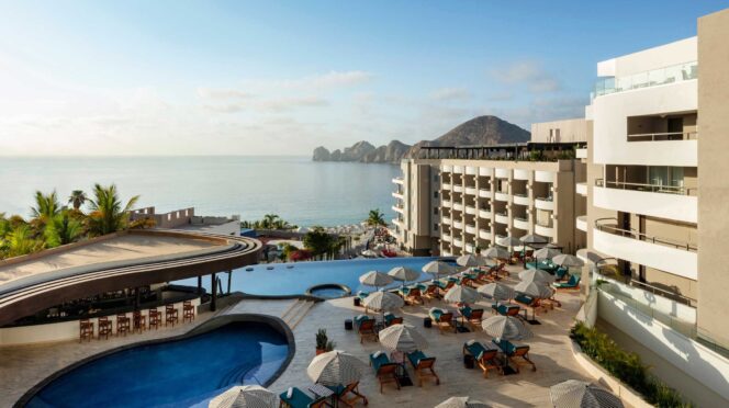 Resort pool area with numerous lounge chairs and striped umbrellas, ocean view in the background under a clear sky.