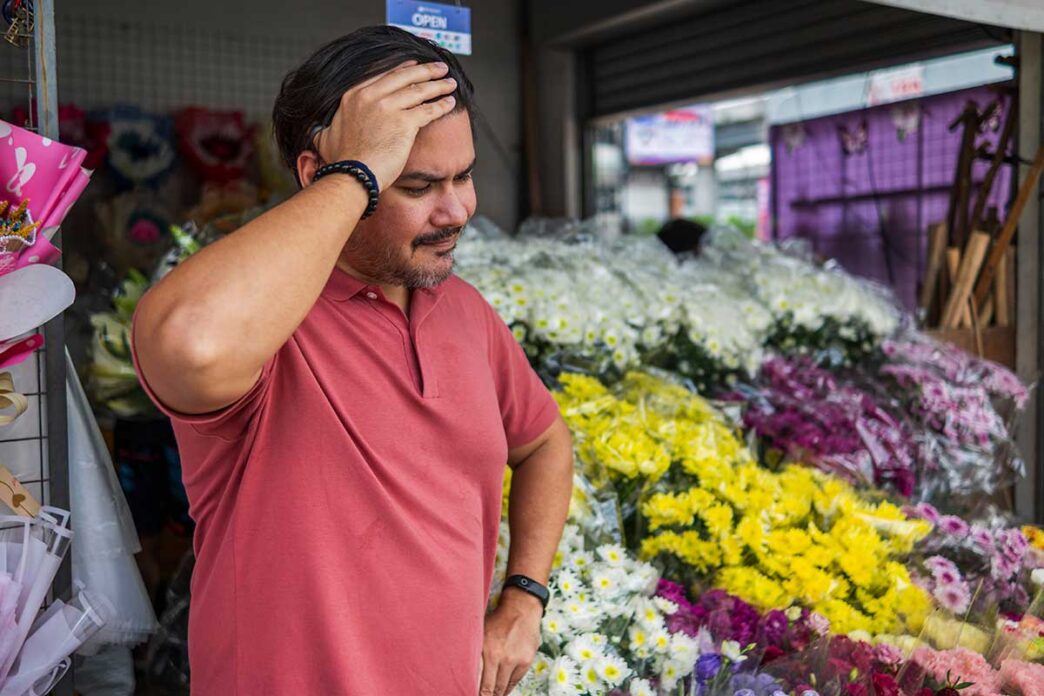 decliningSales - Gringo Gazette Man in a red polo shirt stands in a flower shop, hand on his forehead, looking pensive among colorful bouquets.