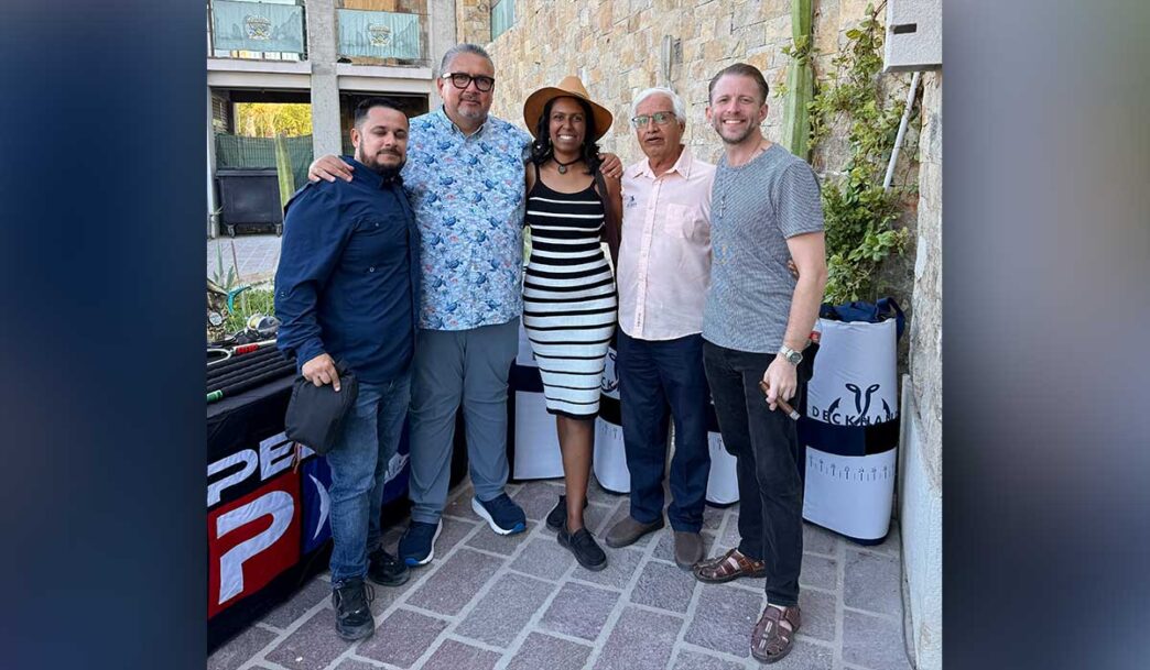Five adults posing together outdoors, smiling for a group photo in front of a stone wall and a table with banners (left-to-right: man in navy shirt, man in floral shirt, woman in striped dress and hat, older man in pink shirt, man in gray t-shirt).