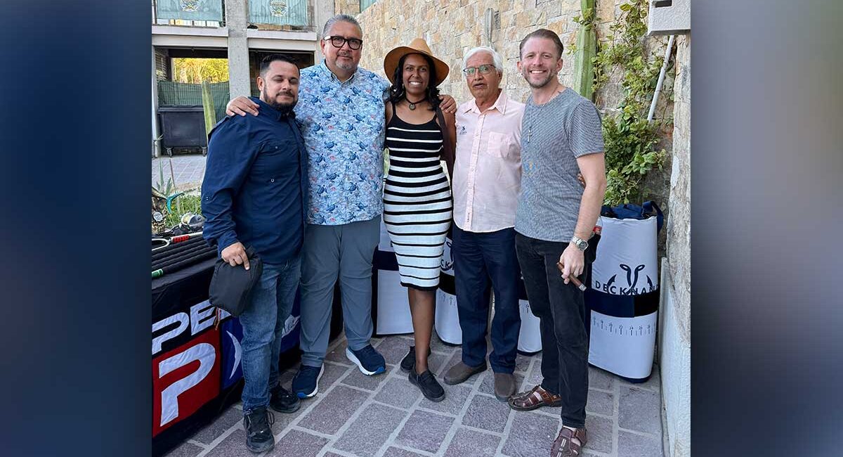 Five adults posing together outdoors, smiling for a group photo in front of a stone wall and a table with banners (left-to-right: man in navy shirt, man in floral shirt, woman in striped dress and hat, older man in pink shirt, man in gray t-shirt).