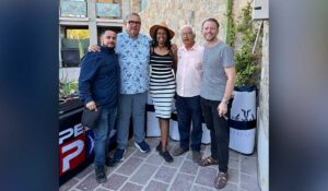 Five adults posing together outdoors, smiling for a group photo in front of a stone wall and a table with banners (left-to-right: man in navy shirt, man in floral shirt, woman in striped dress and hat, older man in pink shirt, man in gray t-shirt).