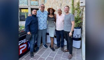 Five adults posing together outdoors, smiling for a group photo in front of a stone wall and a table with banners (left-to-right: man in navy shirt, man in floral shirt, woman in striped dress and hat, older man in pink shirt, man in gray t-shirt).