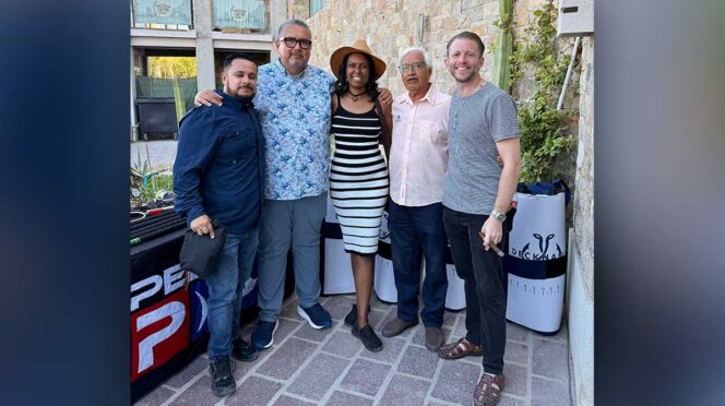 Five adults posing together outdoors, smiling for a group photo in front of a stone wall and a table with banners (left-to-right: man in navy shirt, man in floral shirt, woman in striped dress and hat, older man in pink shirt, man in gray t-shirt).