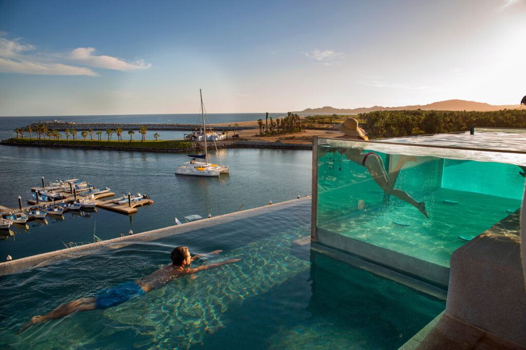 poolside sessions ganzo - Gringo Gazette Swimming pool with clear water overlooks a marina and calm sea at sunset; person partially visible in the pool near a glass barrier.