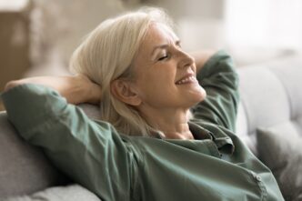 Older woman with gray hair reclining on a sofa, eyes closed and a gentle smile on her face.