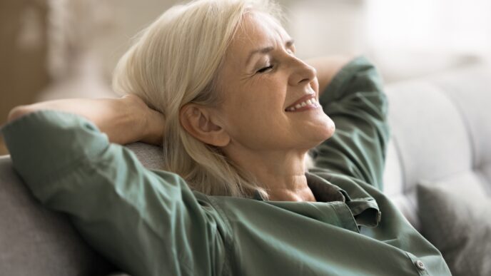 Older woman with gray hair reclining on a sofa, eyes closed and a gentle smile on her face.