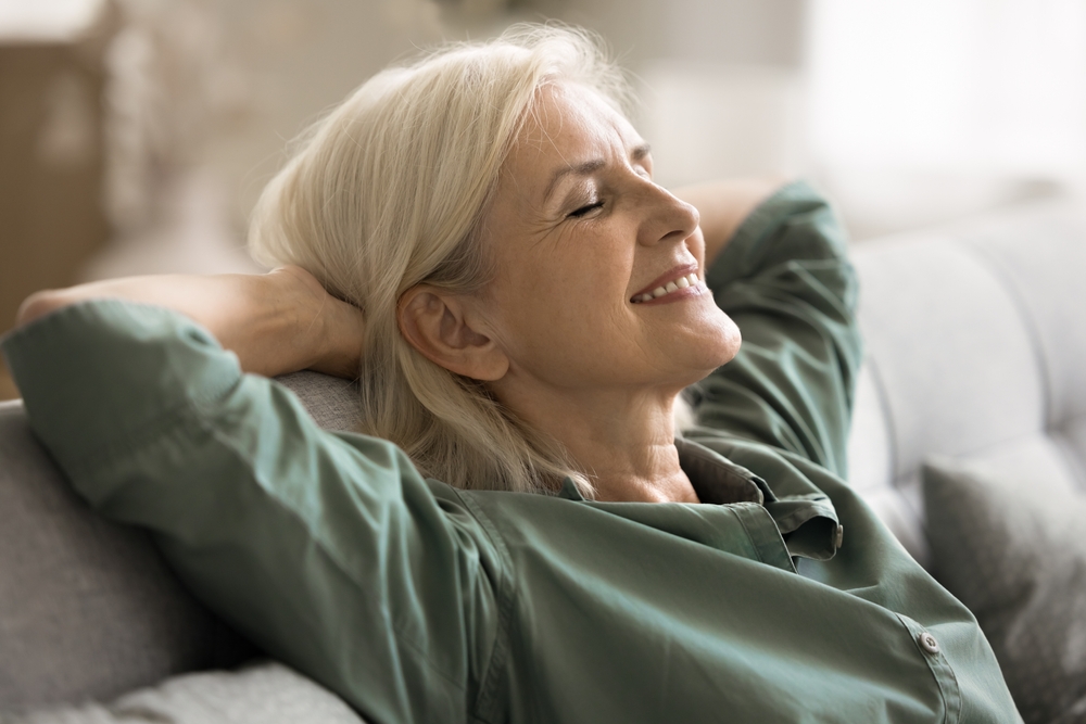 Older woman with gray hair reclining on a sofa, eyes closed and a gentle smile on her face.