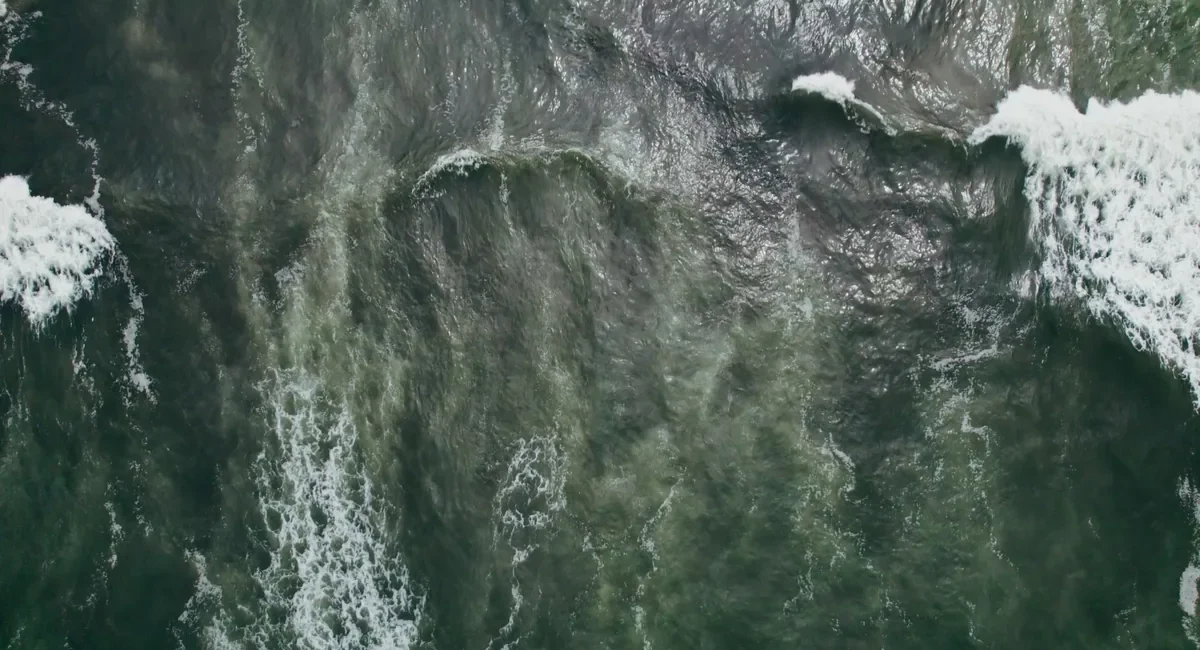 Aerial view of dark green ocean with breaking waves and white foam along the edges, showing swirling water patterns.