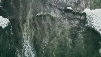 Aerial view of dark green ocean with breaking waves and white foam along the edges, showing swirling water patterns.