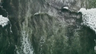 Aerial view of dark green ocean with breaking waves and white foam along the edges, showing swirling water patterns.