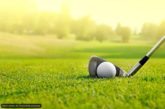 Golf ball resting beside a golf club on a sunlit green fairway with a blurred background.