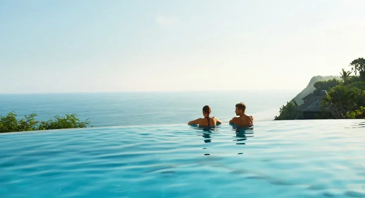 Two people sit in an infinity pool, gazing at the calm blue ocean and distant coastline with tropical greenery in the background.