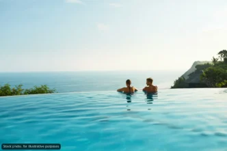 Two people sit in an infinity pool, gazing at the calm blue ocean and distant coastline with tropical greenery in the background.