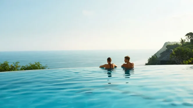 Two people sit in an infinity pool, gazing at the calm blue ocean and distant coastline with tropical greenery in the background.