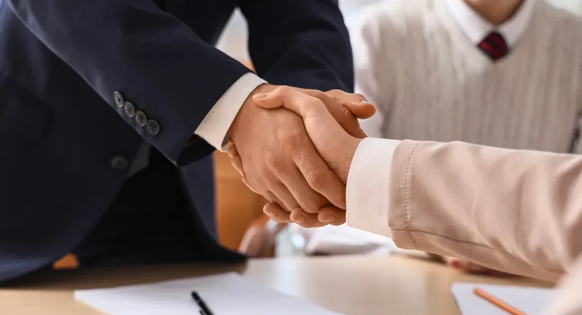 Two professionals shake hands over a desk after signing, with papers and a pen visible on the table.