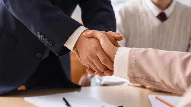 Two professionals shake hands over a desk after signing, with papers and a pen visible on the table.