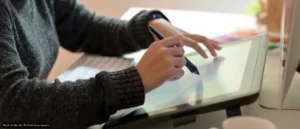 Person in a dark sweater drawing on a graphic tablet with a stylus at a desk.