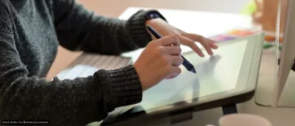 Person in a dark sweater drawing on a graphic tablet with a stylus at a desk.