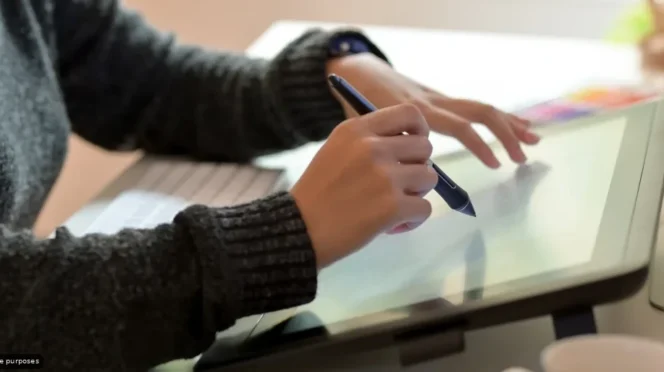 Person in a dark sweater drawing on a graphic tablet with a stylus at a desk.
