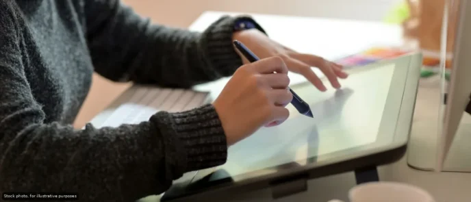 Person in a dark sweater drawing on a graphic tablet with a stylus at a desk.