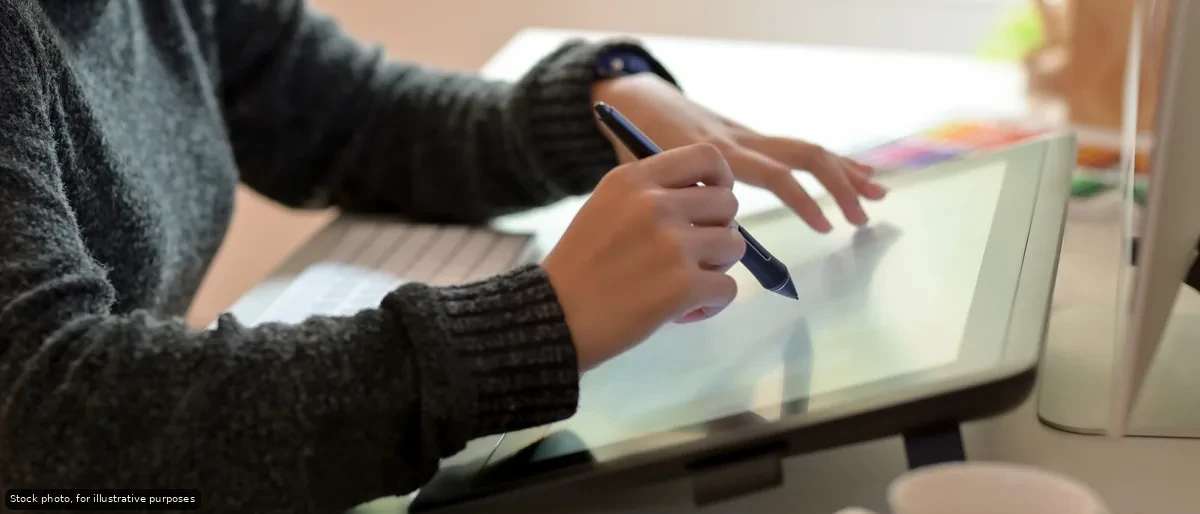 Person in a dark sweater drawing on a graphic tablet with a stylus at a desk.