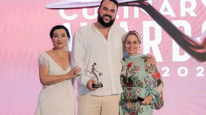 Three people on a stage posing with an award; man in a white shirt holds a trophy, flanked by two women in a white dress and a floral dress, pink backdrop.