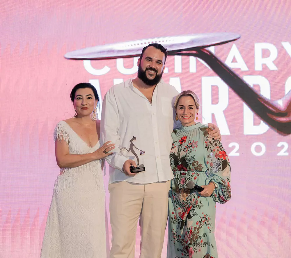 Three people on a stage posing with an award; man in a white shirt holds a trophy, flanked by two women in a white dress and a floral dress, pink backdrop.