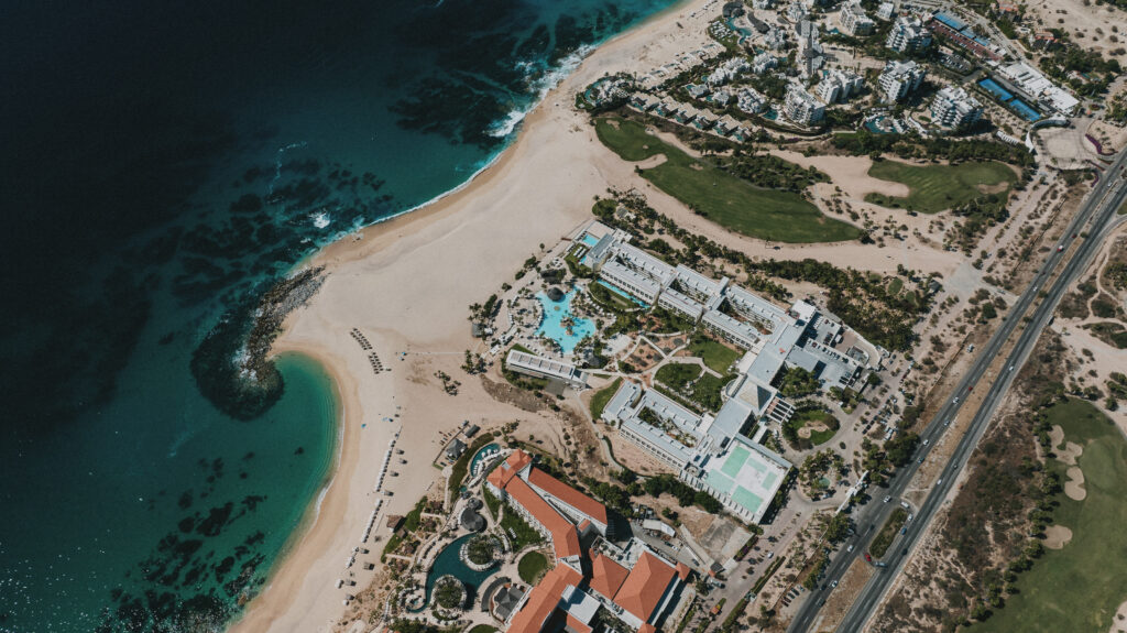 Aerial view of a coastal resort: sandy beach, turquoise water, and cluster of white hotel buildings along a sea-front road with a golf greens layout nearby.
