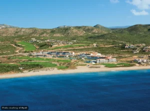 A beachfront resort with white buildings, green lawns, and a blue sea under a clear sky (aerial view).