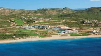 A beachfront resort with white buildings, green lawns, and a blue sea under a clear sky (aerial view).