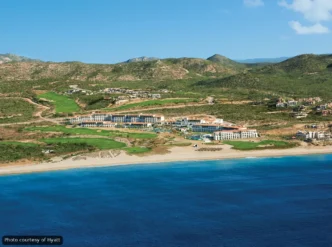 A beachfront resort with white buildings, green lawns, and a blue sea under a clear sky (aerial view).