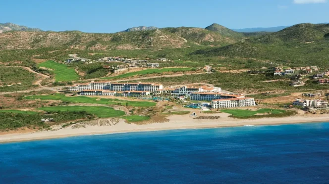 A beachfront resort with white buildings, green lawns, and a blue sea under a clear sky (aerial view).