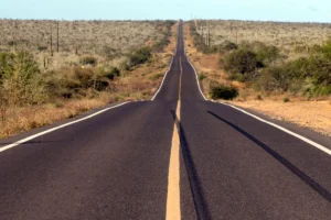 Paved desert highway extending straight into the horizon, flanked by scrubby vegetation and low hills.