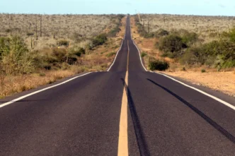 Paved desert highway extending straight into the horizon, flanked by scrubby vegetation and low hills.