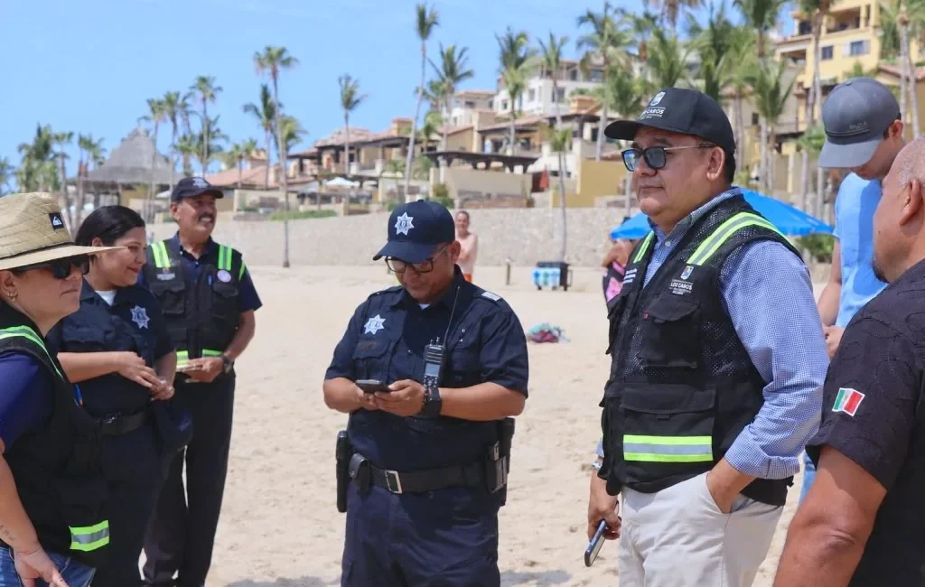 Group of police officers in navy uniforms on a sunny beach, some checking devices, with palm trees and resort buildings in the background.