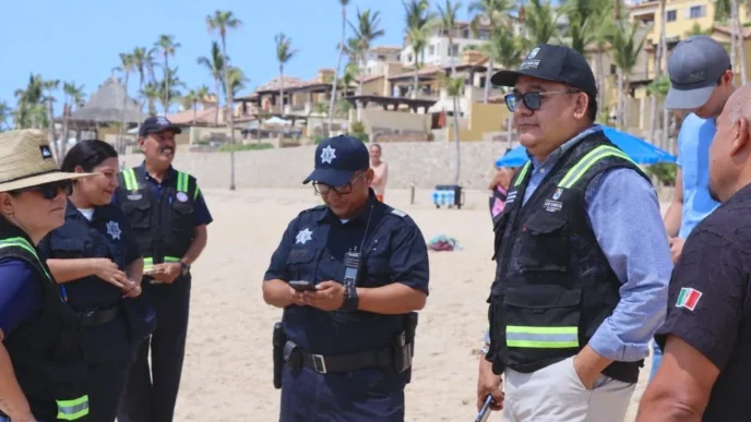 Group of police officers in navy uniforms on a sunny beach, some checking devices, with palm trees and resort buildings in the background.