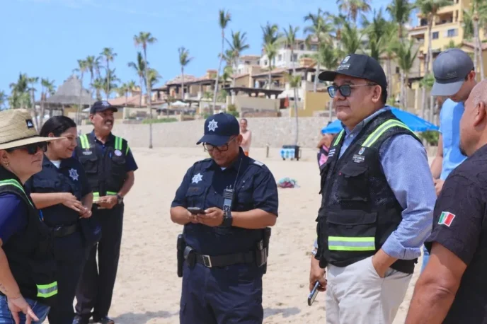 Group of police officers in navy uniforms on a sunny beach, some checking devices, with palm trees and resort buildings in the background.