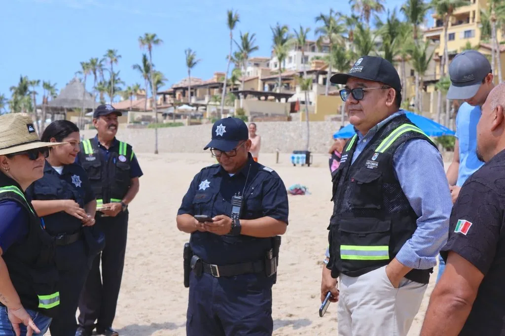 Los Cabos Guarantees Emergency Access at El Médano Beach - Gringo Gazette Group of police officers in navy uniforms on a sunny beach, some checking devices, with palm trees and resort buildings in the background.