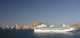 Large white cruise ship sailing near jagged rocky islets off a calm blue sea under a clear sky.