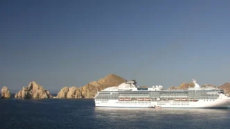 Large white cruise ship sailing near jagged rocky islets off a calm blue sea under a clear sky.