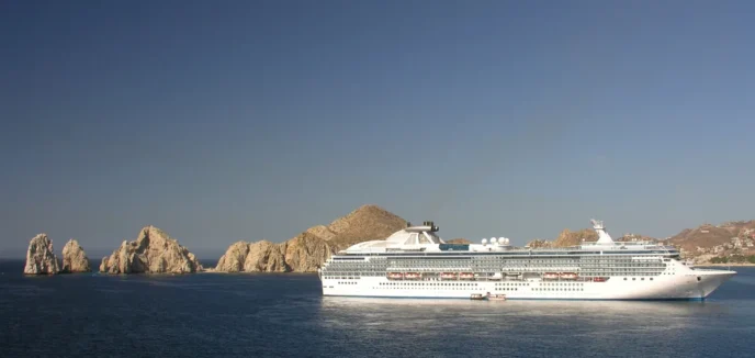 Large white cruise ship sailing near jagged rocky islets off a calm blue sea under a clear sky.
