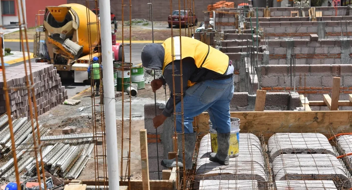 Construction worker in a yellow safety vest bends over to secure steel rebar for a concrete foundation at a construction site, with a cement mixer and blocks in the background.