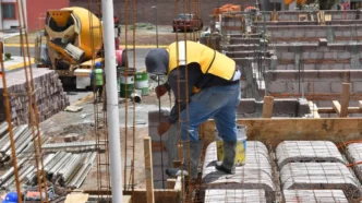 Construction worker in a yellow safety vest bends over to secure steel rebar for a concrete foundation at a construction site, with a cement mixer and blocks in the background.