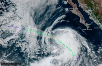 Satellite image of a hurricane off the western Mexican coast near Baja California, with the storm’s eye and spiral clouds visible and a colored forecast track overlay in purple, yellow, and green lines. Alternate: Storm with clear eye approaching Mexico’s Pacific coast; forecast path shown by multi-color lines.