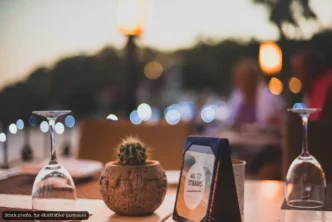 Table setting at a restaurant with a small cactus in a clay pot, a framed 'No To Straws' sign, and two inverted wine glasses; warm bokeh lights in the background.